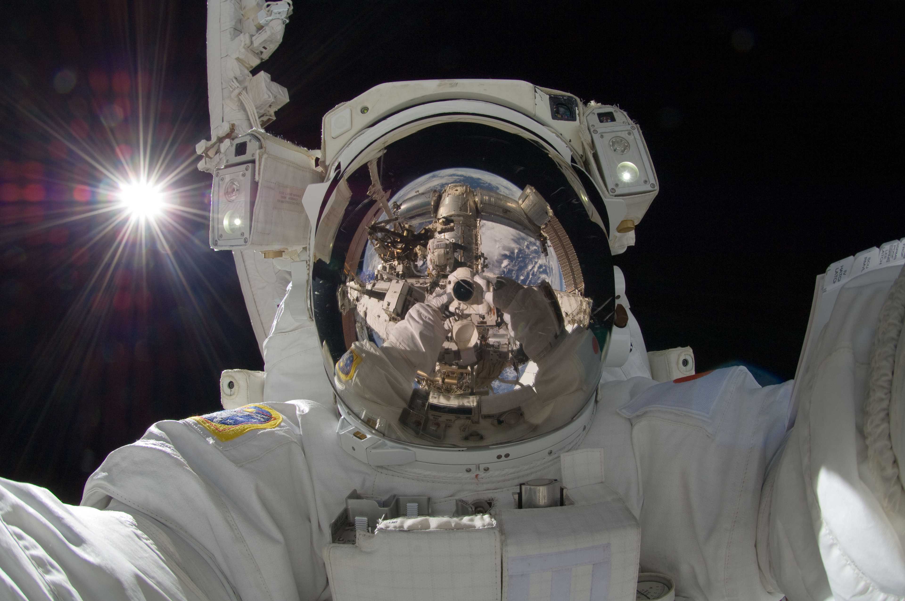 astronaut Aki Hoshide taking a self portrait in a spacesuit with the sun behind him and the camera, ISS and Earth visible in his visor