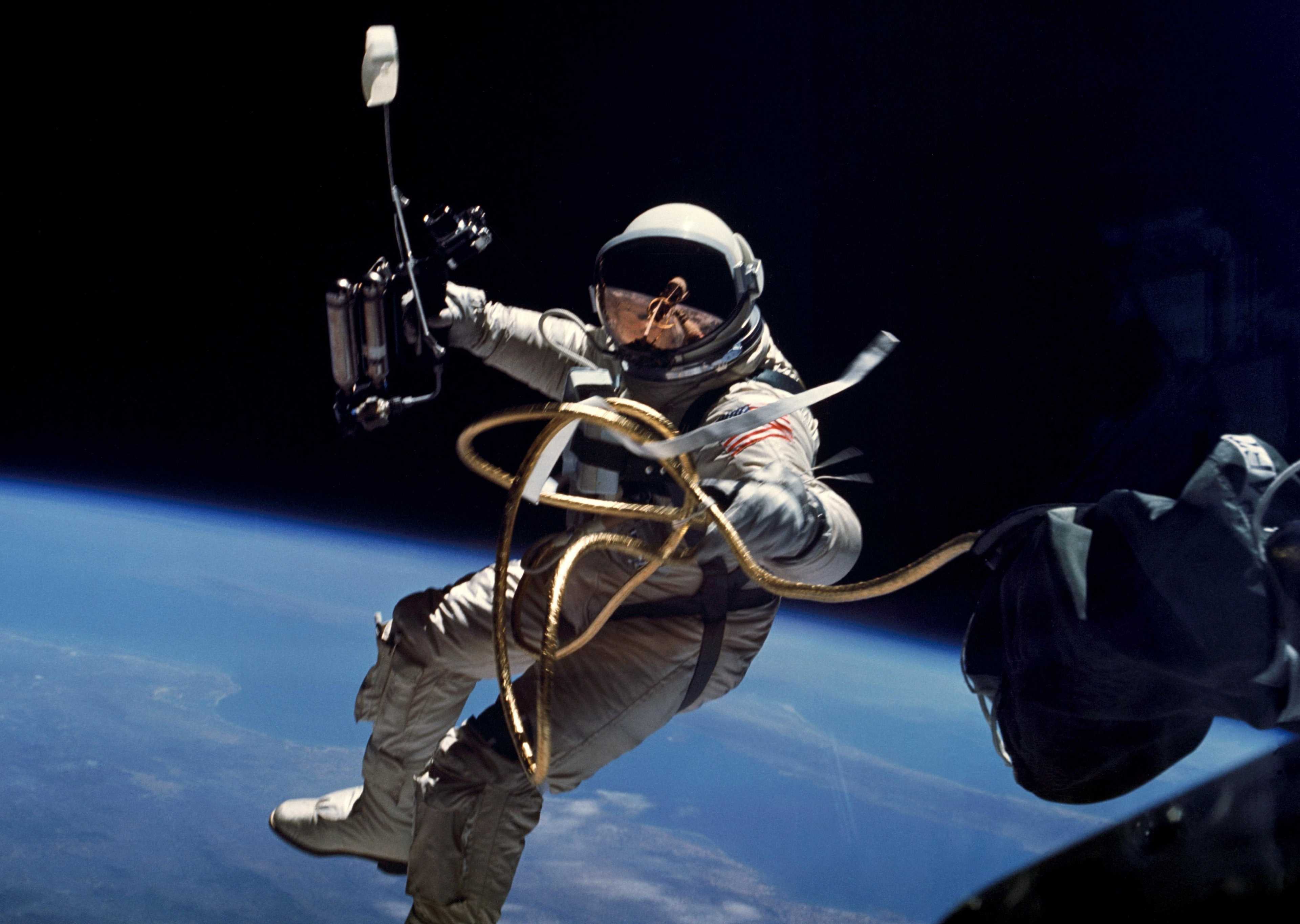 astronaut Edward White in space with the earth below him. a gold tether keeps him attached to Gemini 4.