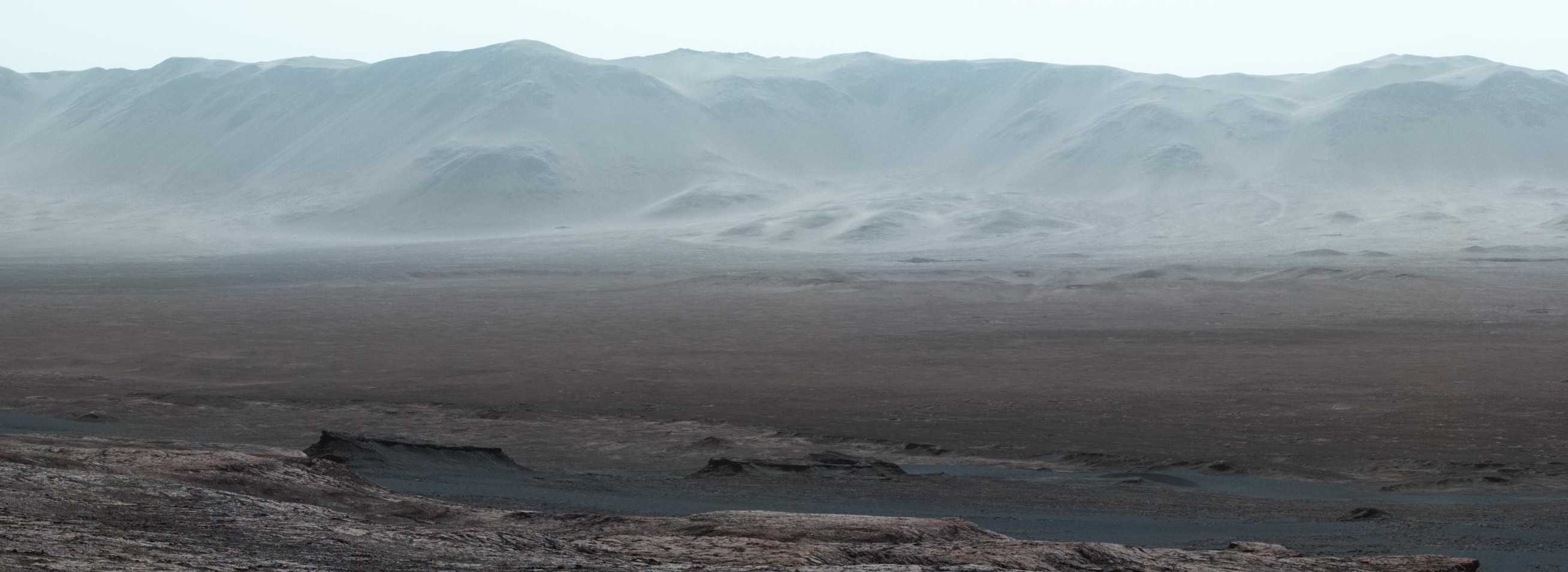 a panorama on mars. the sky is tinted blue, a drop into a valley is in the foreground, with the rising edge of a crater in the background