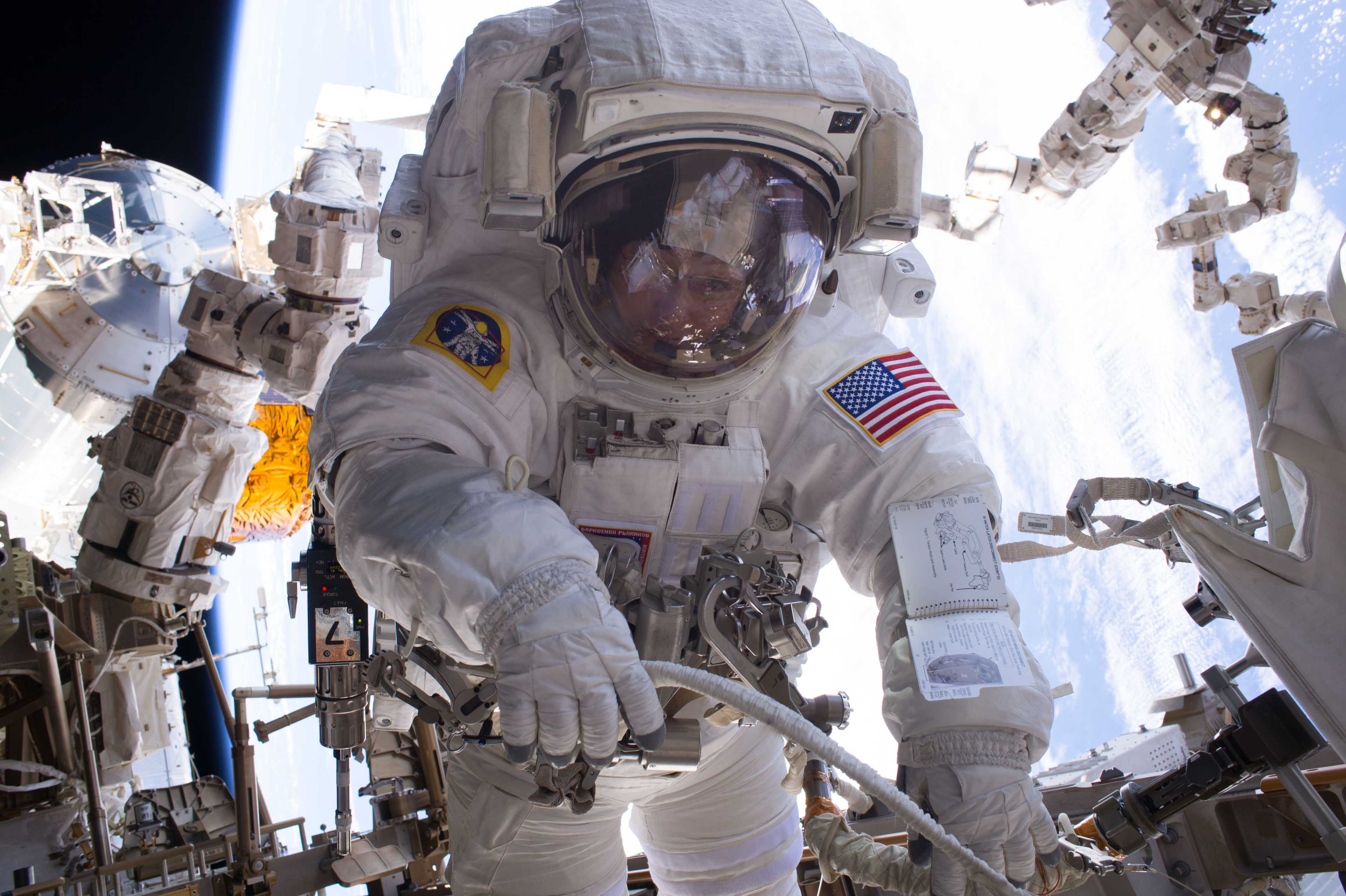 astronaut peggy whitson with ISS equipment behind her, including Canadarm 2. she's holding what looks like tubing outside ISS. her face is actually visible through the visor